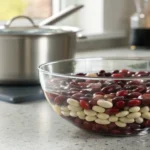 A glass bowl of kidney, black, and white beans soaking in clear water on a modern kitchen counter with a stainless steel pot in the background and natural light streaming through a window.